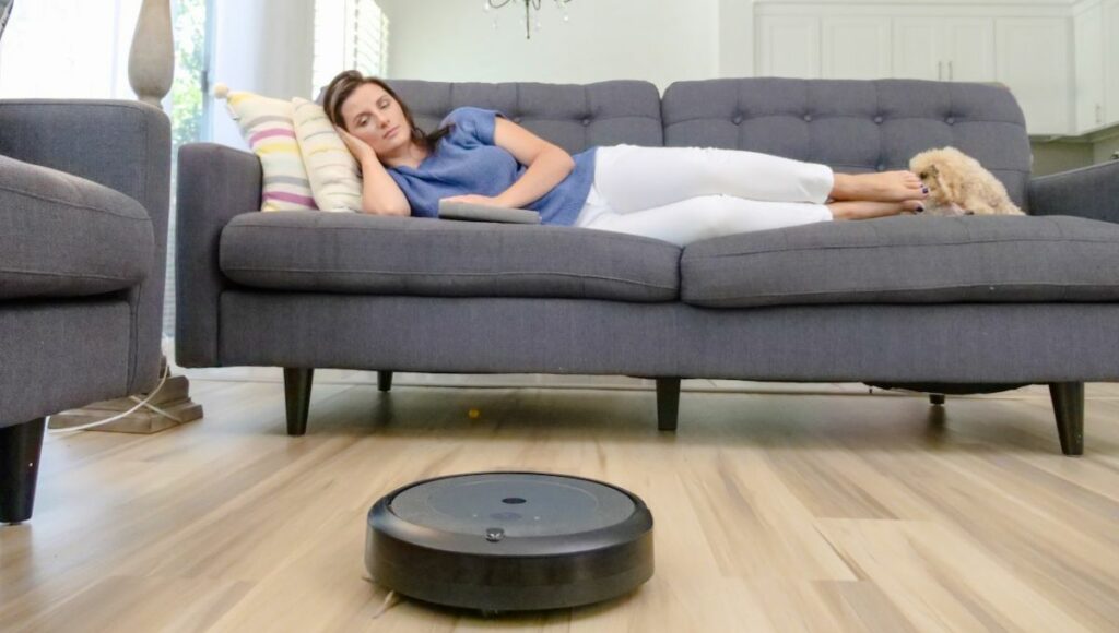 A robot vacuum doing its work with a lady laying on a couch sleeping